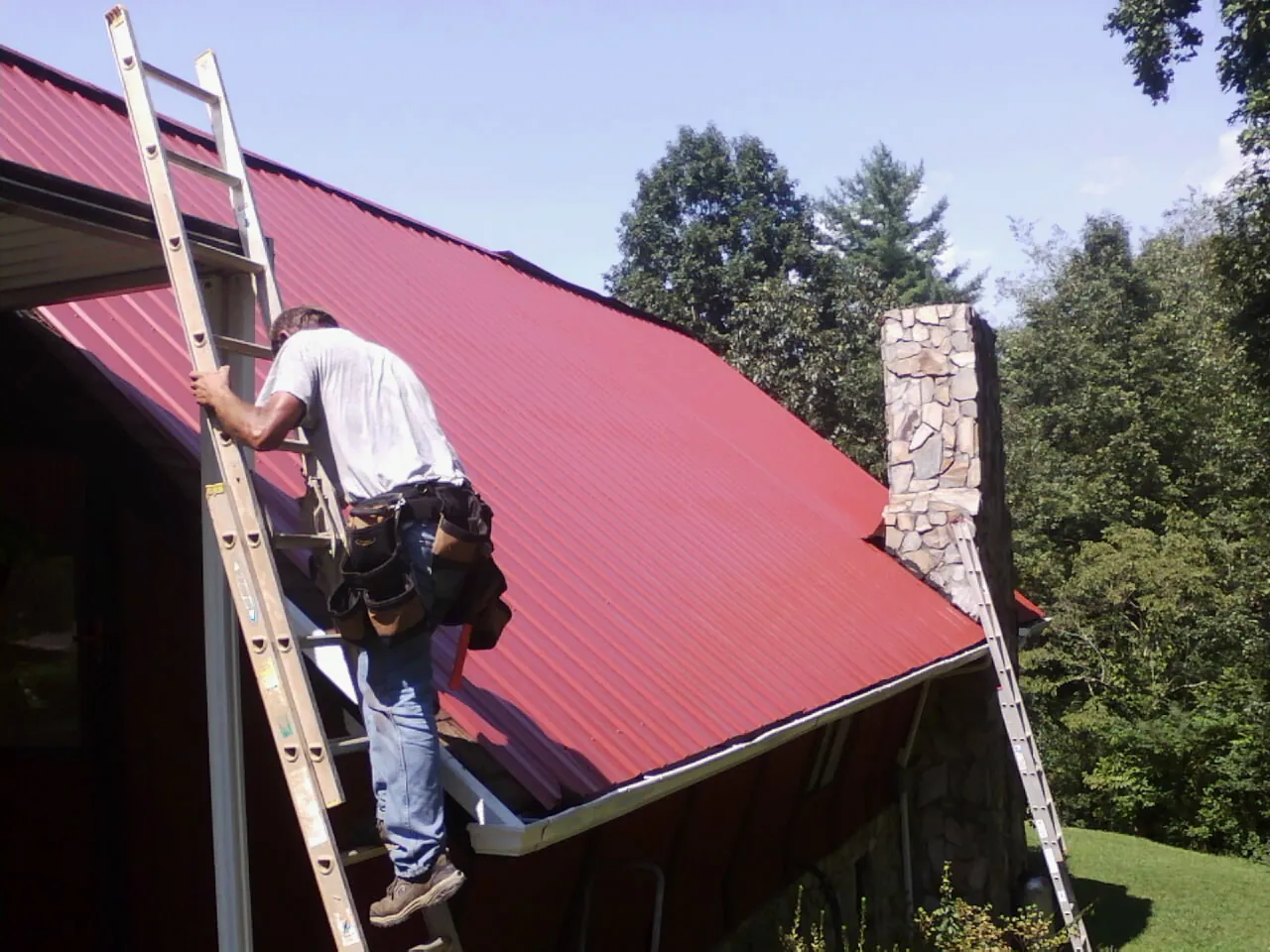 Roofer on a ladder beside a red metal roof and stone chimney — Soffit & Fascia Repair by Roofing Brothers in Lenox Dale